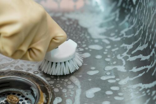 Close-up of a hand in gloves scrubbing a soapy kitchen sink with a brush.