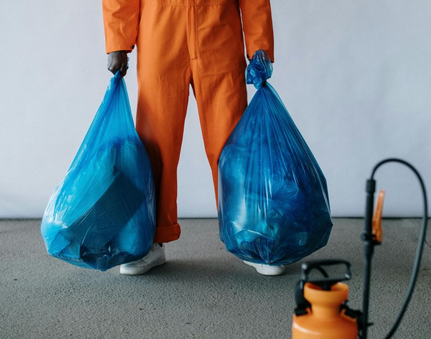 Sanitation worker in orange overalls holding blue trash bags indoors for waste management.